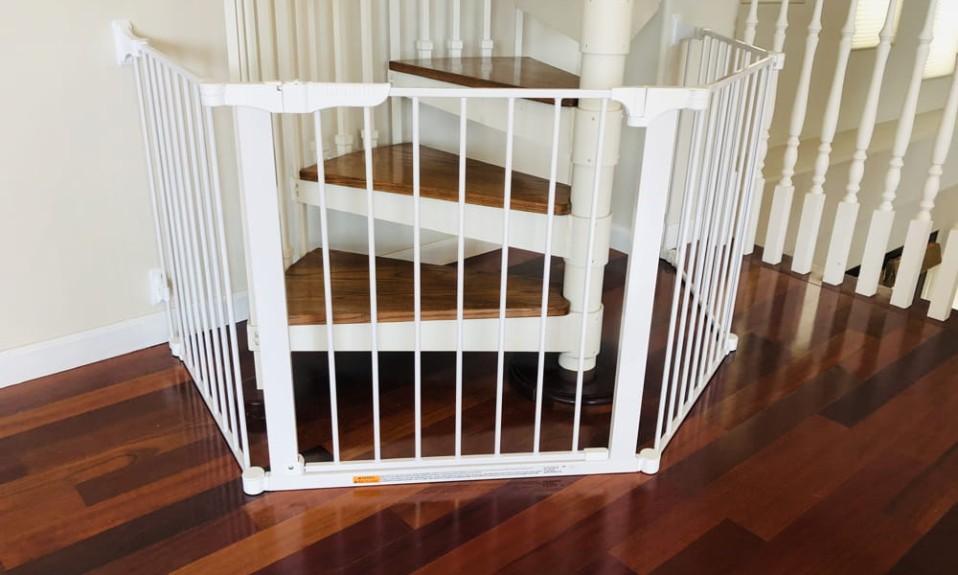 A wooden spiral staircase with a baby gate installed at the top. The gate is made of white metal with vertical slats and a locking mechanism. A baby's hand is reaching through the slats of the gate.